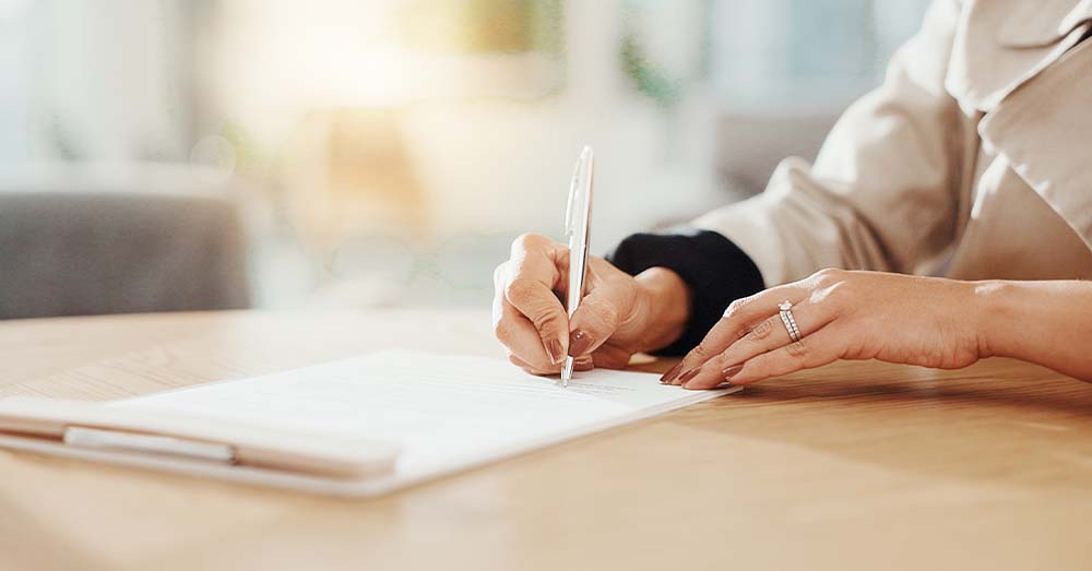Mano que sostiene un bolígrafo plateado y escribe sobre una hoja sobre una mesa.  