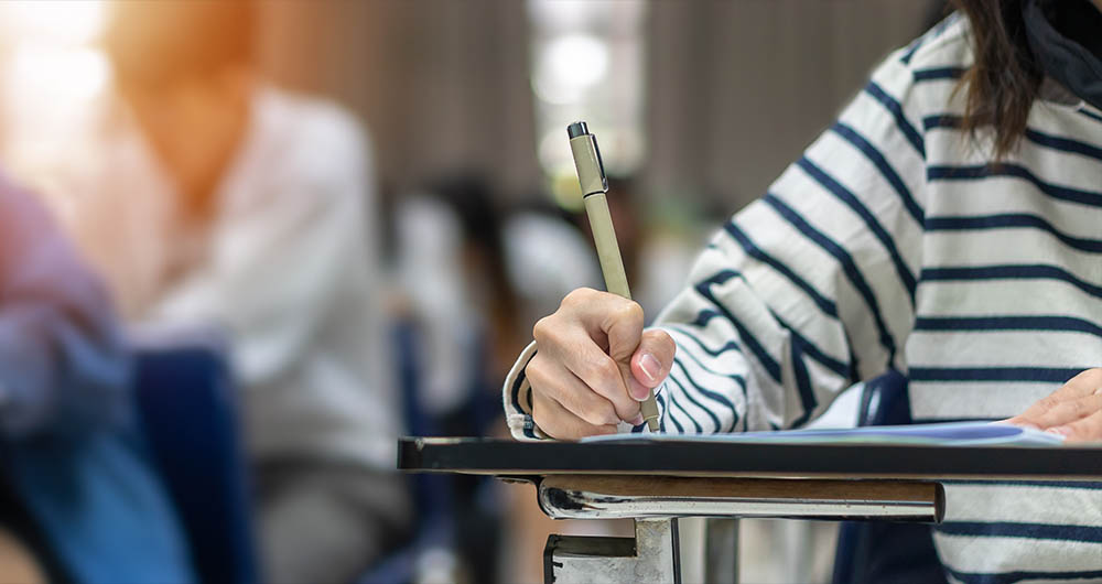 Foto de un pupitre y una estudiante sentada escribiendo en un papel